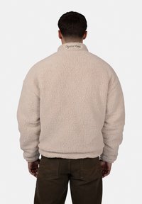 Beiger Fleece-Pullover mit strukturiertem Äußeren, hohem Kragen und "Crystal Paris" auf der Rückseite gestickt. Lockerer Schnitt mit elastischen Bündchen.