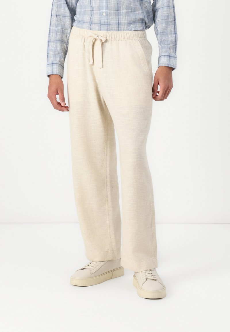 Beige Leinenhose mit lockerer Passform und verstellbarem Kordelzugbund. Mit seitlichen Taschen und strukturiertem Finish. Kombiniert mit weißen Sneakers.