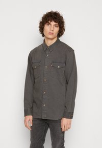 Camisa de mezclilla gris con mangas largas, cierre de botones en la parte delantera, dos bolsillos en el pecho y un discreto logo de la marca. Cuenta con un cuello clásico y botones oscuros.