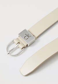 Ceinture en cuir beige avec une boucle en argent poli arborant le logo "ck". Texture lisse et pointe effilée, avec des détails de couture visibles.