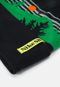 Gorro de malha preto com padrão gráfico verde e cinza, apresentando um sol e árvores. Inclui uma etiqueta do logótipo amarelo da Nike SB na borda.