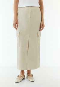 Beige cargo skirt with front slit, featuring two patch pockets on the sides. Smooth, textured fabric with a straight cut design.