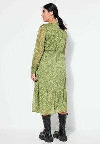 Groene jurk met paisley-patroon, lange doorschijnende mouwen, elastische taille en gelaagde rok. Draagt zwarte combatboots, vanaf de achterkant gezien.