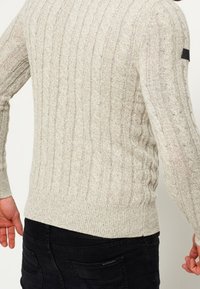 Jasnoszary sweter z dzianiny o fakturze kablowej, z prążkowanym wzorem, dopasowany, z małym czarnym akcentem na ramieniu, noszony z ciemnymi spodniami.