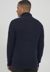 Maglione lavorato a maglia blu navy con colletto alto, polsini e orlo a coste. Presenta un logo sulla spalla sinistra e un motivo a punto testurizzato.
