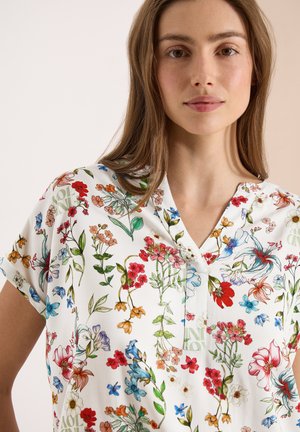 Žena, která má na sobě bílou blůzu s barevným květinovým potiskem obsahujícím červené, modré, oranžové a zelené květiny a listy, stojí na jednobarevném pozadí.