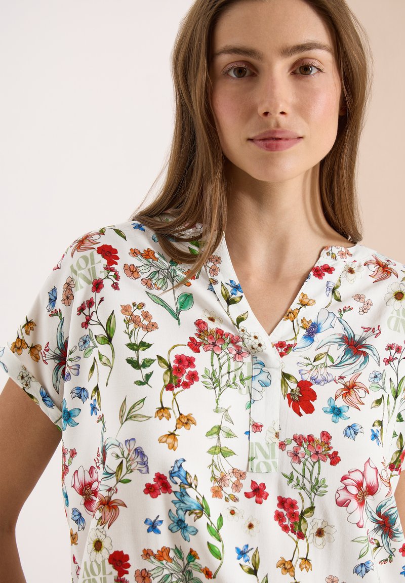 Femme portant un chemisier blanc avec un imprimé floral coloré comprenant des fleurs et des feuilles rouges, bleues, orange et vertes, debout sur un fond uni.