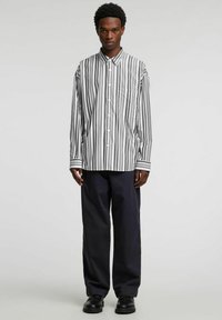 Czarna i biała koszula w paski z długim rękawem, kołnierzykiem, całkowitym zapięciem na guziki i pojedynczą kieszenią na piersi. Noszona z granatowymi spodniami i czarnymi butami.