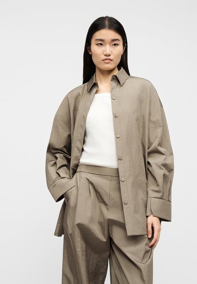 Γυναίκα που φοράει άνετο πουκάμισο σε χρώμα ταουπέ με κουμπιά και παντελόνι φαρδύ σετ με το πουκάμισο, από πάνω λευκό αμάνικο τοπ, στέκεται με τα χέρια στις τσέπες.