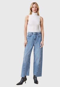 Top ajustado de cuello alto blanco, jeans de pierna ancha azul claro con costuras frontales, combinados con botas negras de tobillo y postura recta contra un fondo neutro.