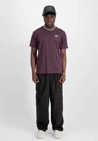 T-shirt crew neck bordeaux avec un petit logo, associé à un pantalon cargo noir. Le modèle porte une casquette noire et des chaussures beiges, se tenant devant un fond blanc.