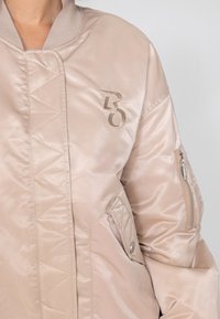 Padded bomber jacket w jasnym beżu, z teksturowaną kieszenią na zamek, diamentowym przeszyciem oraz wyraźnym haftowanym logo na piersi.