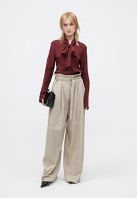 Burgunderfarbene Seidenbluse mit Schleifchen-Detail, kombiniert mit hoch taillierten, weit geschnittenen beigen Hosen. Das Model hält eine kleine schwarze Handtasche.