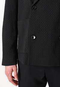 Schwarze zweireihige Jacke mit strukturiertem Muster. Mit schwarzen Knöpfen und einer Seitentasche ausgestattet, kombiniert mit schmal geschnittenen schwarzen Hosen.