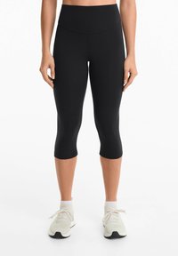 Černé capri legíny s vysokým pasem, vyrobené ze strečového materiálu. Mají hladkou texturu a přiléhavý střih. Nosič má na sobě světlé sneakers.
