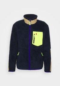 Marinblå fleecejacka med en khakifärgad krage, neongrön bröstficka, två sidofickor och kontrasterande lila dragkedjedetaljer.