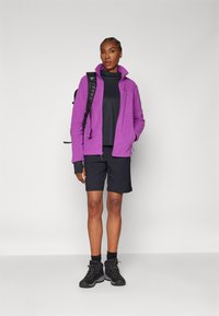 Lila Fleecejacke über einem schwarzen Langarmshirt, kombiniert mit schwarzen Shorts und Wanderschuhen. Das Outfit hat eine strukturierte Oberfläche.
