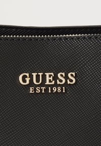 Borsa nera testurizzata con logo dorato "GUESS EST 1981", chiusura con zip nella parte superiore e superficie liscia.