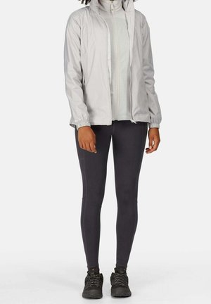 Personne portant une veste zippée gris clair, des leggings noirs ajustés et des chaussures de randonnée noires, debout devant un fond blanc uni.