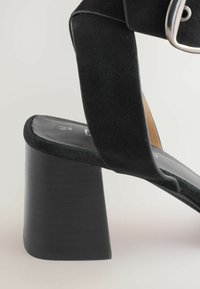 Black heeled sandal featuring a wide strap and block heel on a neutral background, emphasizing the shoe’s texture and design details.