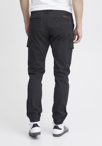 Uomo che indossa pantaloni cargo neri con polsini elasticizzati e sneaker bianche, in piedi con la schiena rivolta verso un sfondo bianco.