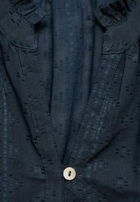 Mörkblå skjorta i tyg med en subtil texturerad mönster. Har en volangkrage och en enda knappstängning. Ljust färgad knappdetalj.