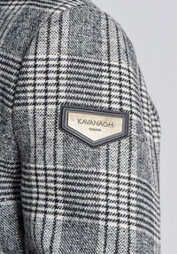 Šedá a bílá kostkovaná látka s hladkým povrchem. Obsahuje černé a zlaté logo Kavanagh Gianni na rameni.