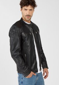 Schwarze Lederjacke mit Reißverschluss, zwei Brusttaschen und gepolsterten Schultern. Getragen über einem weißen Hemd mit blauen Jeans.