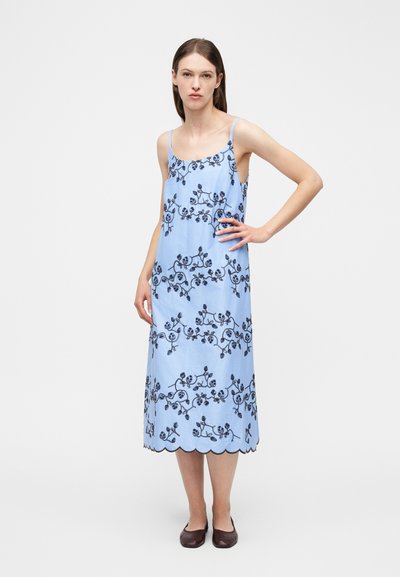 Γυναίκα που φοράει ανοιχτό μπλε midi φόρεμα με μαύρη κεντημένη ανθοδέσμη, οδοντωτό τελείωμα, λεπτές τιράντες και σκούρα επίπεδα παπούτσια, στέκεται με το χέρι στη μέση.