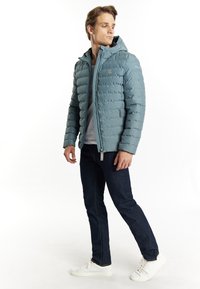 Ljusblå pufferjacka med en quiltad design, dragkedjestängning och huva. Bärs över en grå skjorta, tillsammans med mörka jeans och vita sneakers.