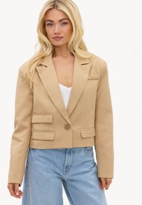 Femme blonde portant un blazer court beige avec des poches à rabat et un jean taille haute bleu clair, debout devant un fond uni.