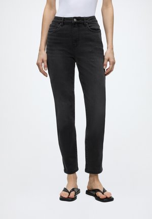 Person iført højtaljede sorte jeans med lige ben, hvid tætsiddende ærmeløs top og sorte sandaler med åben tå på en ensfarvet baggrund.