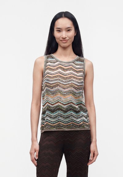 Femme aux longs cheveux noirs portant un haut sans manches à motif zigzag multicolore et un pantalon foncé avec une texture zigzag.