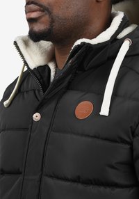 Zwarte donzen jas met een gestructureerde stof, witte imitatiebont voering, leren logopatch en contrasterende witte trekkoordaccenten.