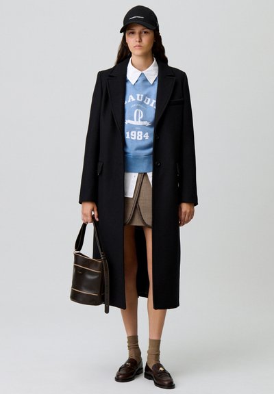 Cappotto lungo nero su un maglione blu con grafica, colletto di camicia bianco, gonna a quadri, borsa marrone, calze marroni e mocassini.
