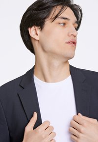 Blazer blu navy scuro con vestibilità slim, caratterizzato da un rever a lancia e tessuto leggermente strutturato. Indossato sopra una maglietta bianca semplice.