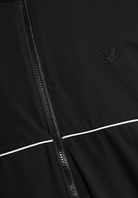 Μαύρο μπουφάν με κεντρικό μαύρο φερμουάρ, λεπτομέρεια λευκής επένδυσης κοντά στο κάτω μέρος και σκούρο λογότυπο αετού στη δεξιά πλευρά.