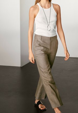 Femme portant un haut blanc sans manches, un pantalon beige taille haute, des sandales noires et un long collier pendentif, marchant à l’intérieur sur un sol sombre.