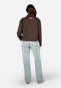 Pruun puuvillane jope punase logo applikatsiooniga selja ülaosas, kombineeritud helesiniste laia säärega teksadega, nähtud tagant.