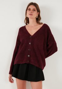 Burgundi színű, puha fleece kardigán V-nyakkal, hosszú ujjakkal és hat nagy gombbal az elején. Fekete szoknyával párosítva.