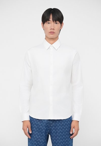 Chemise blanche à manches longues avec un col pointu et une fermeture à boutons sur le devant, en tissu lisse. Associée à un pantalon bleu à motifs.