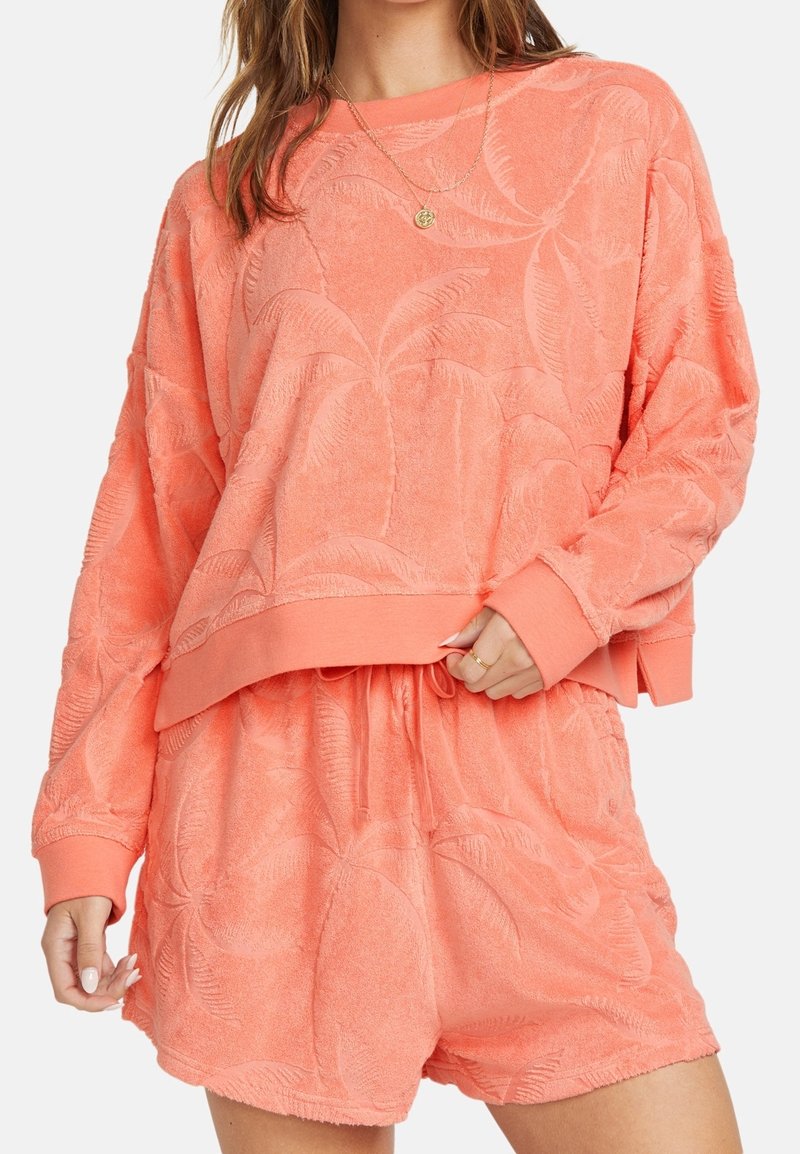 Femme portant un sweat-shirt en éponge texturée corail et un short assorti avec un motif de feuilles de palmier, tenant l'ourlet du sweat-shirt d'une main.