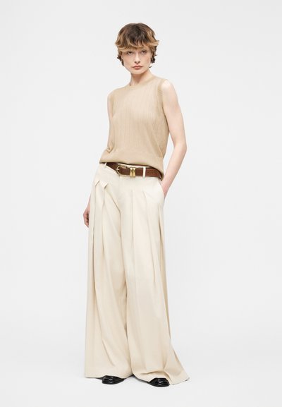 Νεαρό άτομο που φοράει αμάνικη πλεκτή μπλούζα μπεζ, κρεμ παντελόνια με φαρδύ πόδι και ψηλή μέση, καφέ ζώνη και μαύρα παπούτσια, στέκεται με το ένα χέρι στην τσέπη.