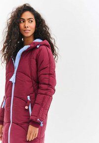 Bordový prošívaný kabát s modrými akcenty, který má kapuci, přední zapínání na zip a dvě boční kapsy. Texturovaný prošívaný design s elastickými manžetami.