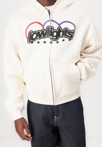 Felpa con zip di colore crema chiaro, con un grande logo nero, anelli rossi, blu e viola, e cinque stelle nere. Raggiunge un look casual e sportivo.
