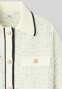 Veste chemise en tweed crème avec des accents noirs. Présente un col pointu, une poche avant et des boutons dorés le long de la patte de boutonnage. Tissu texturé.