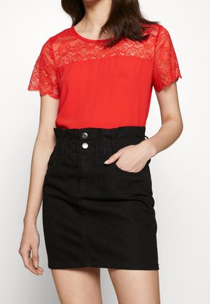 Woman wearing a red lace short-sleeve top tucked into a high-waisted black skirt with buttons and a pocket.