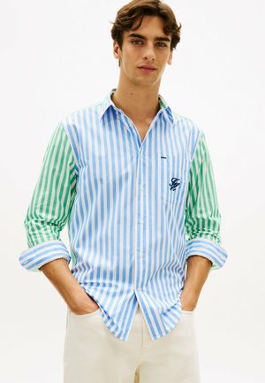 Mann trägt ein langärmliges Hemd mit vertikalen Streifen; der Körper hat blau-weiße Streifen, die Ärmel grün-weiße Streifen.