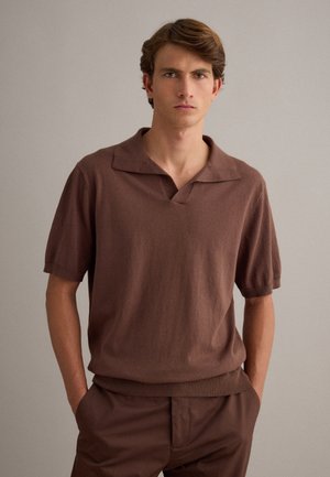 Giovane uomo con capelli castano chiaro che indossa una polo marrone a maglia a maniche corte e pantaloni marroni abbinati su uno sfondo beige uniforme.