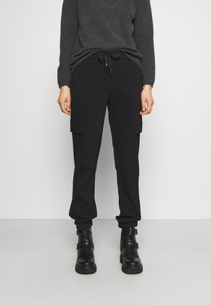 Person iført sorte cargobukser med snøre i taljen, grå sweater og sorte snørestøvler stående mod en ensfarvet baggrund.